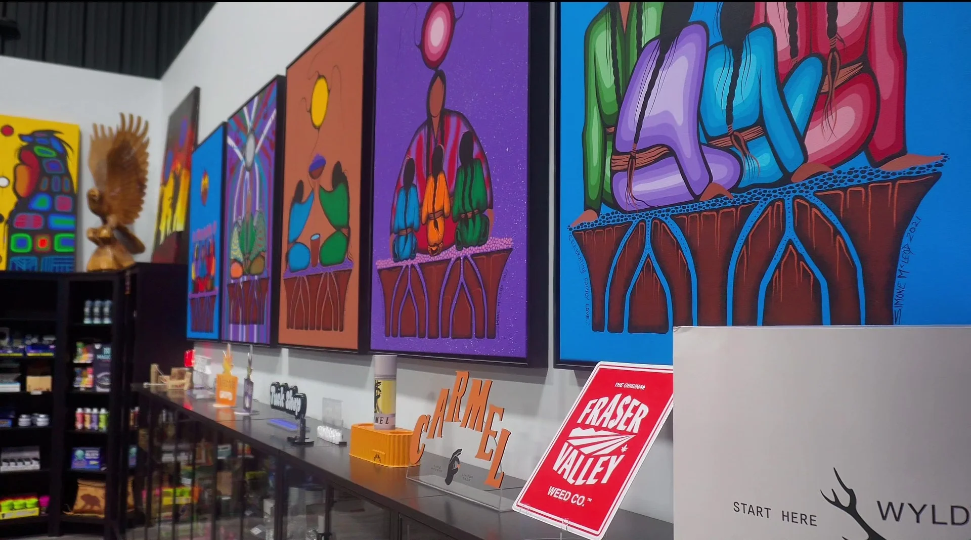 An angled shot showcasing four different Indigenous paintings in a set. There is a wood eagle carving on the top shelf in the back above indistinct cannabis accessories.